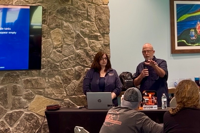 Two presenters stand behind a table with a laptop in a conference room, a stone wall behind them, audience in foreground
